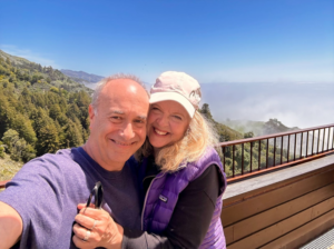 SARK and her husband David in Big Sur, CA on an overlook showing mountain ranges coated in fog beneath a bright blue clear sky